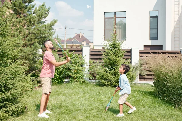 年轻的成年人穿着休闲装 和他的小儿子在后院草坪上打羽毛球 侧视拍摄 — 图库照片