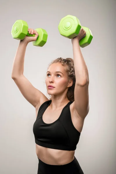 アクティブなブロンド女の子で黒スポーツウェア保持グリーンダンベルで手以上彼女の頭の上に物理的な運動のために灰色の背景 — ストック写真