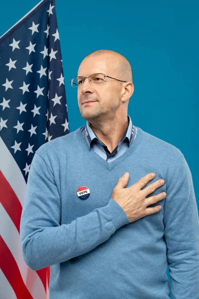 Başarılı bir iş adamı, eli göğsünde kameranın önünde yıldız ve çizgili bayrak ve mavi arka plana karşı duruyor.