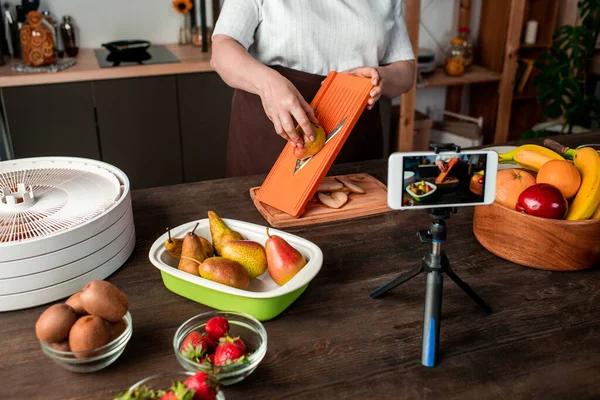 Çağdaş ev hanımı ya da blog yazarı akıllı telefon kamerasının önünde mutfak masasının yanında duruyor ve kuru meyve yapma videosu kaydediyor.