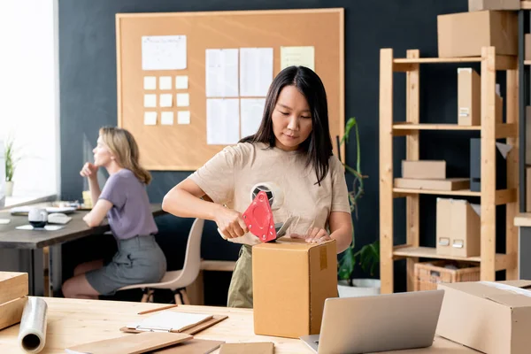 Çevrimiçi atölyede çalışan Asyalı genç bir kadın ya da depo odası karton kutulardan birini seloteyp ile mühürlerken sipariş vererek paketliyor.