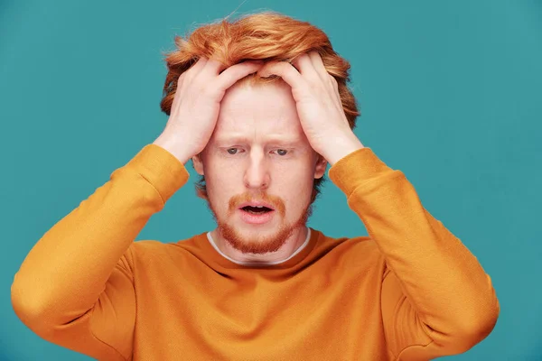 Portrait of shocked young redhead guy with beard keeping mouth open in astonishment against blue background