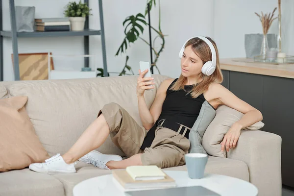 ヘッドフォンのブロンドの女の子とカジュアルスマートフォンの画面を見ながらソファでリラックスして見て何かを選択 — ストック写真