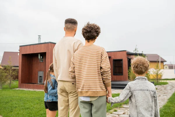 Yeni evin önündeki yeşil çimlerde dikilen güncel aile ve gündelik giysiler içindeki iki küçük çocuğun arka görüntüsü.