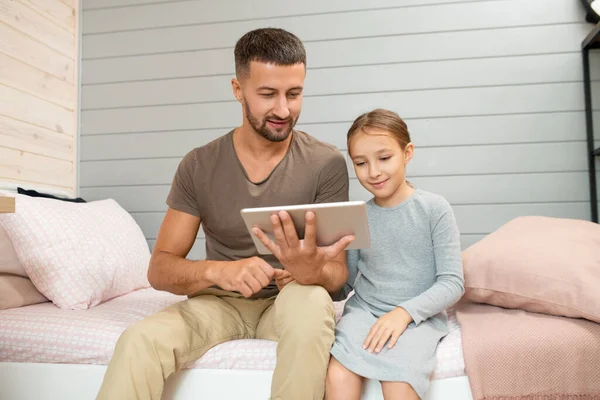 年轻面带微笑的爸爸 带着数码平板电脑坐在他可爱的小女儿旁边 一边看电影 一边翻看新闻 — 图库照片