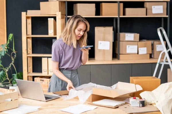 Cep telefonlu genç sarışın kadın siparişi paketleyip bir müşteriye göndermeden önce kutuda katlanmış süveterin fotoğrafını çekiyor.