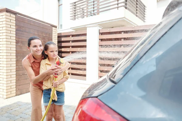 Modern anne ve kızı birlikte dışarıda vakit geçiriyorlar. Hortumla araba yüzeyinden sabunu suyla yıkıyorlar.