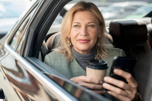 一个成熟的女人坐在出租车后座上 透过开着的窗户 一边喝着咖啡 一边用智能手机打滚 — 图库照片