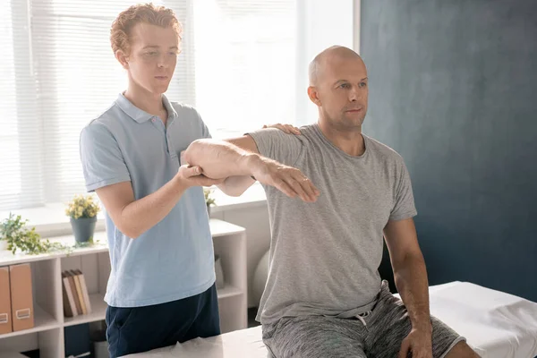 在医务室 年轻的物理治疗师在协助病人进行适当的康复训练的同时 亦支持病人肘部的手臂 — 图库照片