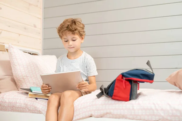 Cute little blond schoolboy with digital tablet sitting on bed and watching online video or movie after school among backpack and books