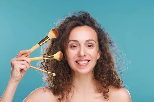 Yüzünde üç kozmetik fırça tutan dişlek bir kadın mavi arka plana profesyonel makyaj uygulayacak.