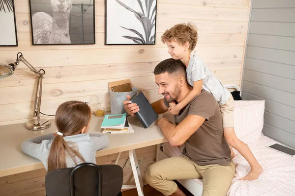 Küçük çocuk, babasının arkasında durup eğlenirken boynuna sarılıyor. Masa başı kızıyla konuşan genç adam.