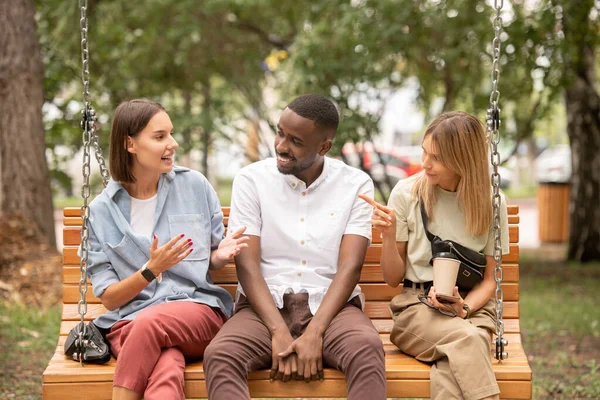 İki kız arasında tahta salıncakta oturan mutlu genç Afrikalı adam tartışırken birine bakıyor.