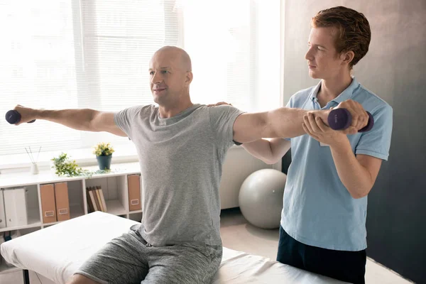 Mavi polo gömlekli genç fizyoterapist klinikte rehabilitasyon sırasında halterli bir hastanın kolunu uzatıyor.