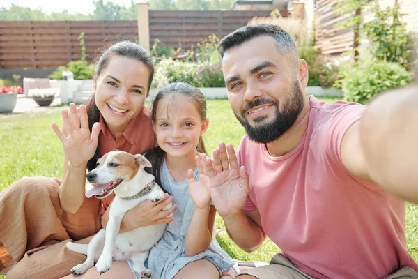 Father taking selfie shot of himself and his family waving hands at camera, girl holding cute dog