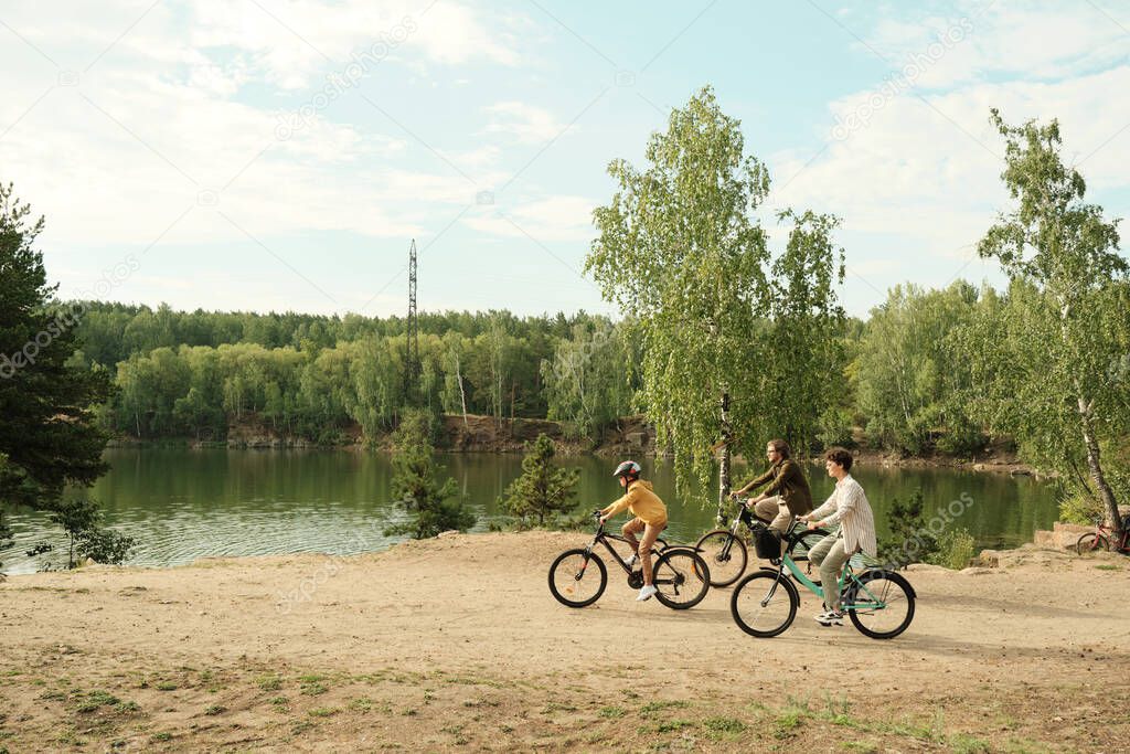 Side view of young family sitting on bicycles and moving along riverbank against green trees while enjoying active rest together