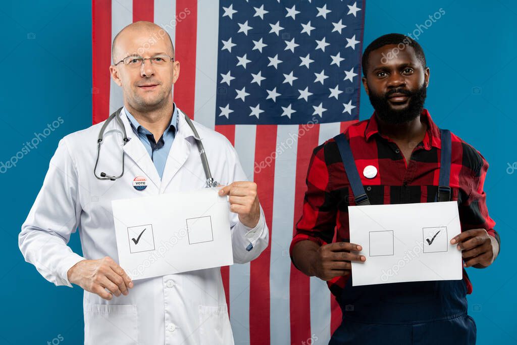 Mature bald doctor and young repairman holding papers with ticks in squares while looking at you against stars-and-stripes flag