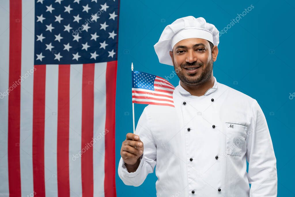 Happy young chef in white uniform holding us flag while standing in front of camera against blue copyspace and stars-and-stripes