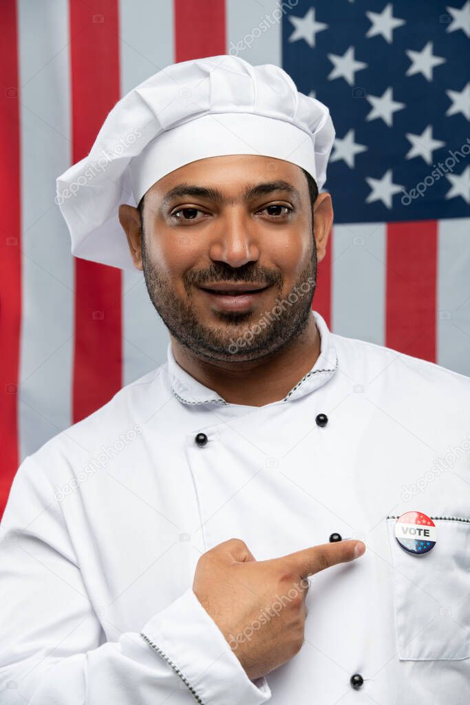 Smiling male chef of restaurant in uniform pointing at vote insignia on chest while standing in front of camera against stars-and-stripes