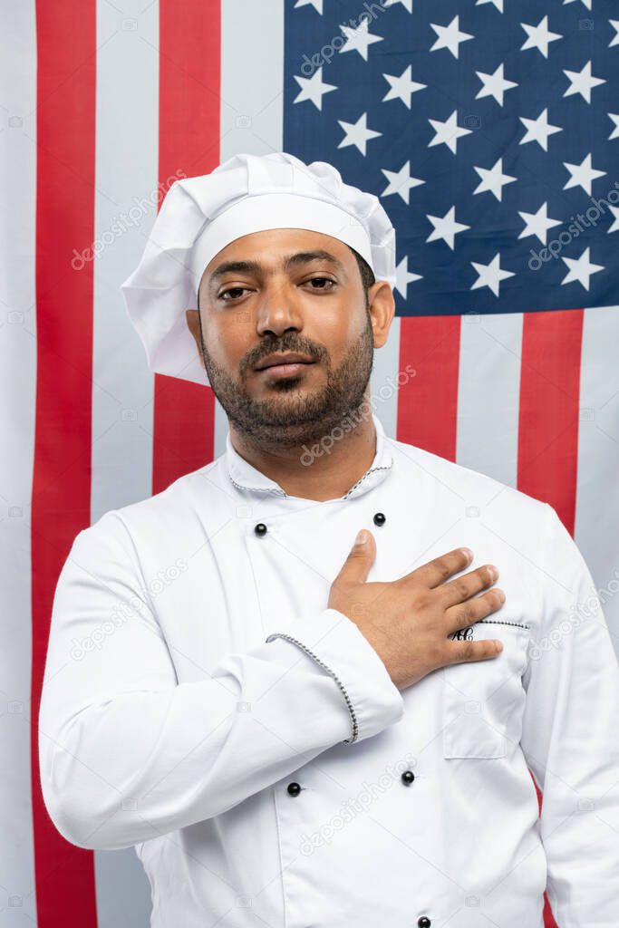 Successful male chef of restaurant with his hand on chest standing in front of camera and looking at you against stars-and-stripes flag