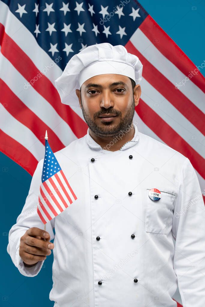 Young serious chef in white uniform holding us flag while standing in front of camera against stars-and-stripes background and looking at you