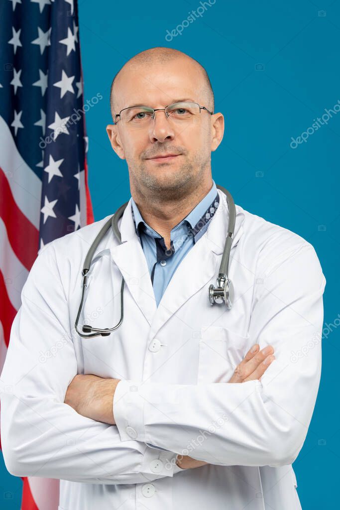 Mature bald clinician in whitecoat crossing arms on chest while looking at you against stars-and-stripes flag and blue background