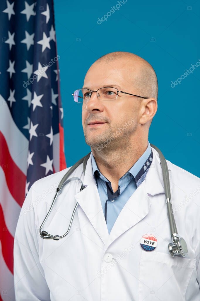 Contemporary bald clinician with vote insignia on whitecoat standing in front of camera against stars-and-stripes and blue background