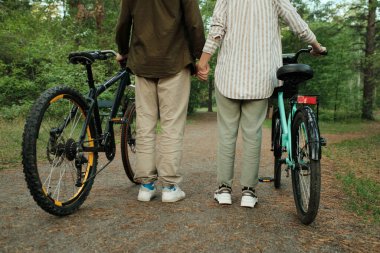 Yeşil ağaçlar arasında kozalaklı iğnelerle kaplı yolda dikilen gündelik kıyafetli genç bisikletçilerin arka görüntüsü.