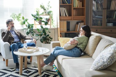 Melankolik genç kadın kanepeye oturmuş yastığına sarılıyor terapi seansı sırasında düşüncelerini psikologlara paylaşıyor.