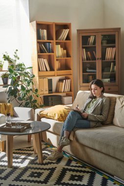 Modern ofis odasındaki rahat koltukta oturan genç bir kadın elinde bir not defteri tutuyor.