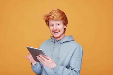 Kapüşonlu genç, kuşkucu, kırmızı sakallı bir adam turuncu arka planda vitamin şişesinin etiketini okuyor.