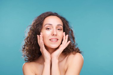 Göz altı bantlı, genç, sağlıklı esmer kadın. Mavi arka planda kameranın önünde derisini yüzerken yüzüne dokunuyor.