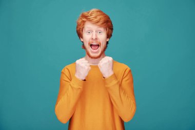 Şaşkın, turuncu kazaklı genç adam mavi arka planda duruyor ve ağzını heyecanla kapatıyor.