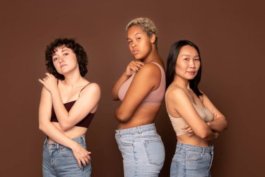Young beautiful intercultural females in tanktops and blue jeans standing in row against brown background in front of camera and looking at you clipart