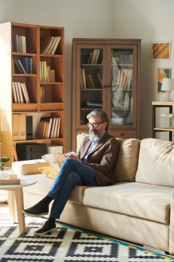 Modern ofis odasındaki kanepede oturmuş notlar alan şık giyimli yakışıklı sakallı adamın dikey fotoğrafı.