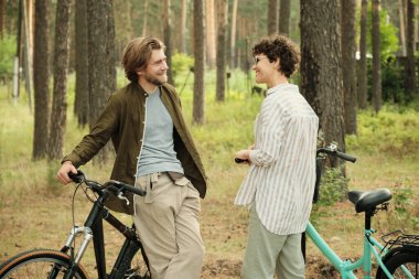 Genç gülümseyen adam güneş gözlüklü güzel kız arkadaşına bakıyor ve yaz tatilinin keyifli anlarını çam ağaçlarının arasında tartışıyor.