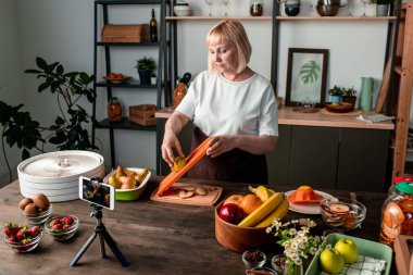 Sarışın, orta yaşlı, akıllı telefon kamerasıyla bakan kadın kış için kurutulmuş meyve yapma videosunu kaydediyor.