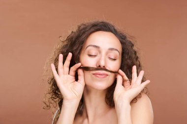 Güzel, genç, gözleri kapalı bir kadın. Bej arka planda dudakları ve burnu arasında bir tutam saç tutarken eğleniyor.