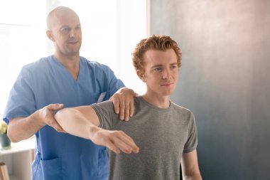 Modern rehabilitasyon kliniklerinde fizyoterapi eğitimi sırasında genç bir hastanın bükülmüş kolunu destekleyen mavi üniformalı yetişkin bir klinisyen.