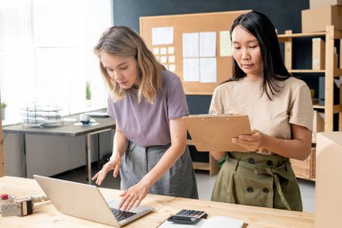 İki kültürlü genç kadın atölyede ya da stüdyoda masanın başında otururken, bir tanesi dizüstü bilgisayar ve meslektaşı not alıyor.