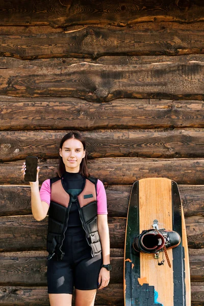 穿着安全夹克和运动服的年轻女冲浪手靠着木制房屋的墙壁站着 用智能手机展示她的自拍 — 图库照片
