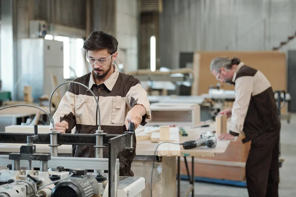 Young serious worker of furniture factory looking at you while standing by workbench on background of workshop interior during work