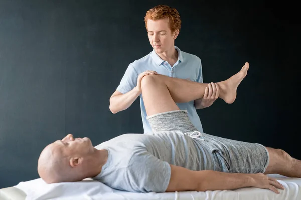 严重的男性理疗师 在康复中心或诊所帮助病人进行体育锻炼时 将病人的腿弯在膝上 — 图库照片