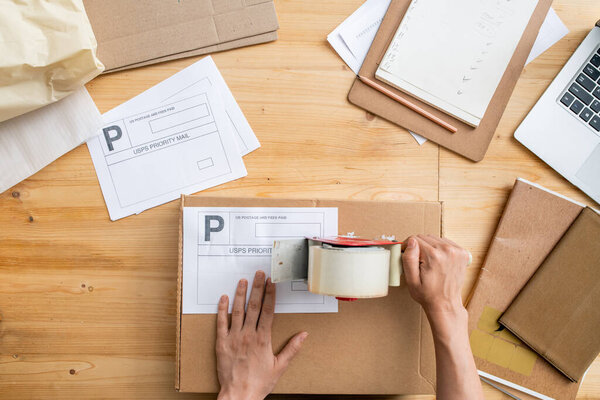 Top view of hand of female manager of online shop with cellotape gun dispenser sealing cardboard box with packed order of client