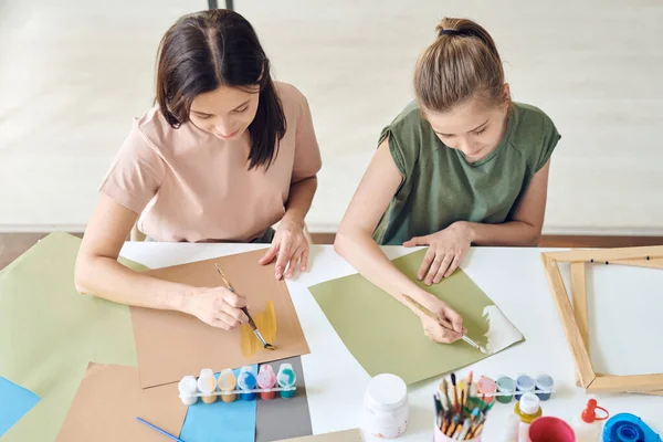 年轻女子和她十几岁的女儿坐在桌旁 手握水彩画或水彩画的画笔 坐在写字台旁 — 图库照片