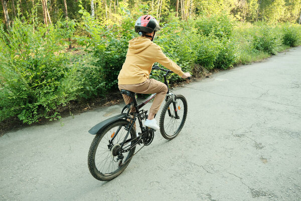 Вернуться к просмотру юношеской езды на велосипеде вдоль дороги против зеленой травы и кустарников во время отдыха в природной среде в выходные дни