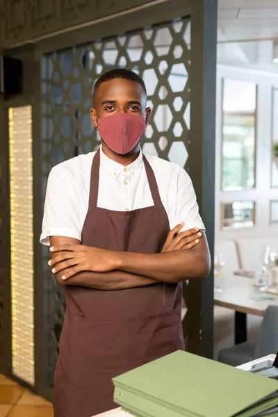 Young contemporary waiter in protective mask and brown apron crossing arms on chest while standing by one of tables in cafe or restaurant