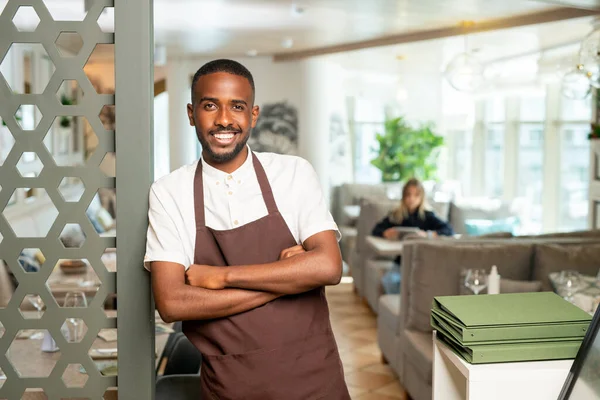 年轻快乐的非洲服务员穿着工作服靠着门在当代餐厅里等新客人 — 图库照片