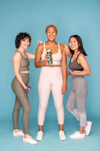 Three young laughing sportswomen in activewear holding bottle of water and dumbbells while going to have training together