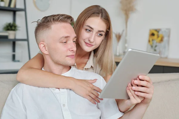 金发碧眼的年轻女子 带着平板电脑拥抱丈夫 弯下腰 在网上商店里给他展示漂亮的衣服或家庭用品 — 图库照片